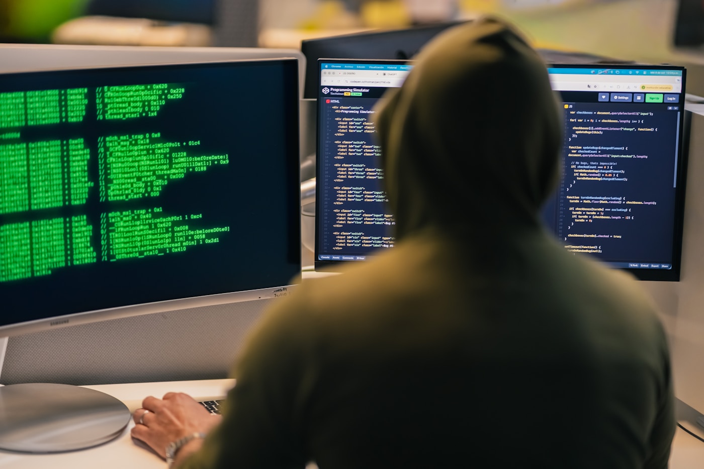 A hooded figure working across multiple computer screens, representing cybersecurity threats
